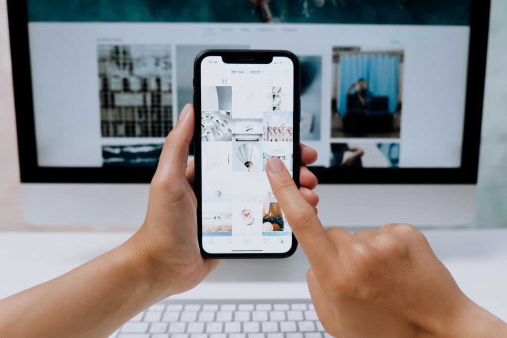 Oseba drži pametni telefon v rokah ter s prstom brska po galeriji slik na ekranu, v ozadju pa je videti računalniški zaslon z odprto spletno stranjo s fotografijami.
